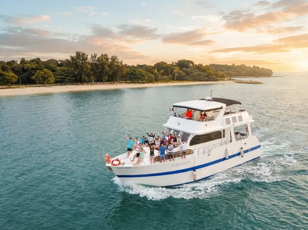 Sardinia Yacht Singapore - A large group of people wave from a white yacht named "SARDINIA" as it cruises on turquoise water near a tropical sandy beach with lush trees, under a vibrant sunset sky at 1 Degree 15 Marin...