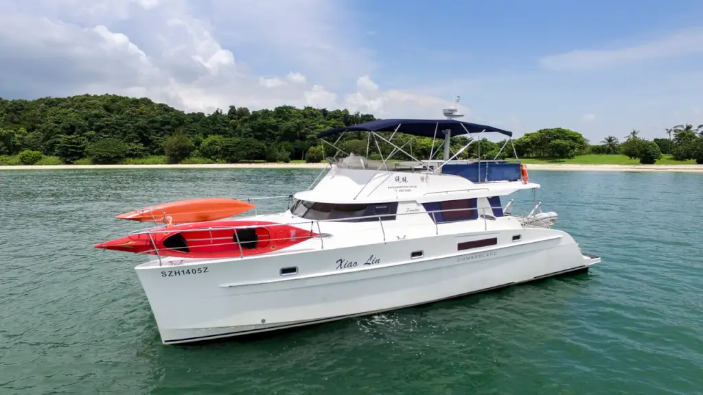 Cumberland 47 - MyEscapade Yacht Charter - A white catamaran yacht with red and orange kayaks on its bow floats on turquoise water near a lush green island with a sandy beach under a partly cloudy sky at Marina at Kep...